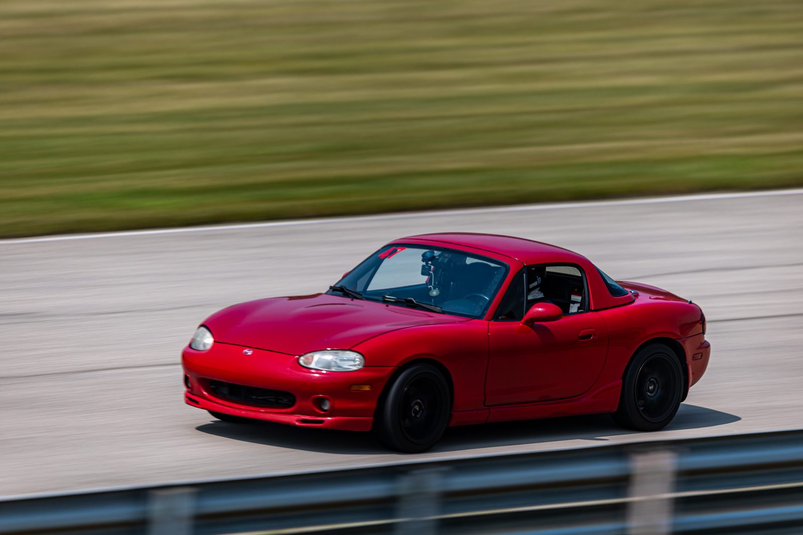 The red Miata side-on at speed on the Autobahn straight, the grass behind it streaked by the camera's pan.