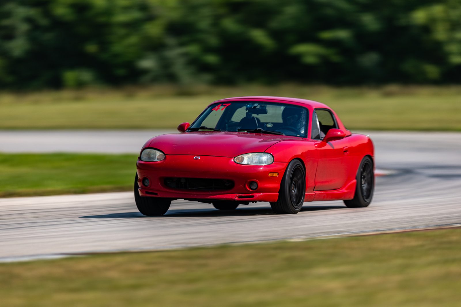 Chuck's red Miata with its hard top on and a number 47 on the windshield, cornering at Autobahn with the trees blurred behind.
