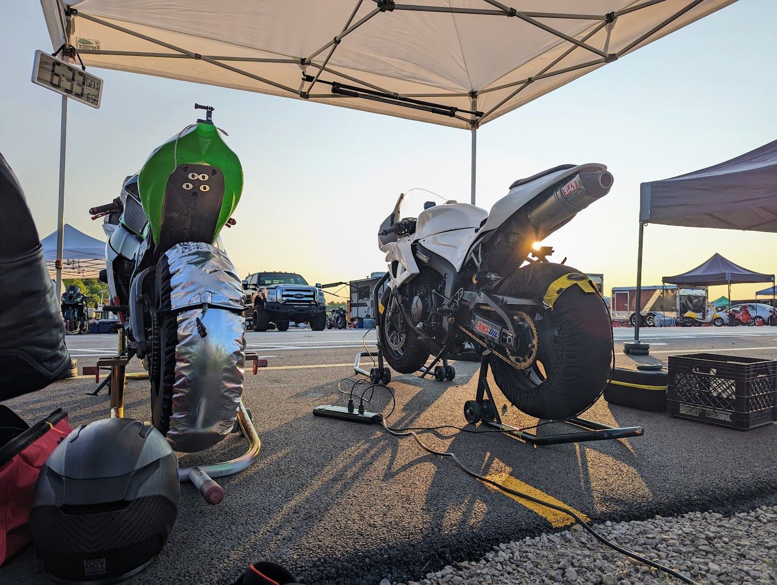 Two race bikes on stands under a canopy in the paddock at sunrise, tire warmers on, and a clock on the canopy leg reading 6:33 SAT.