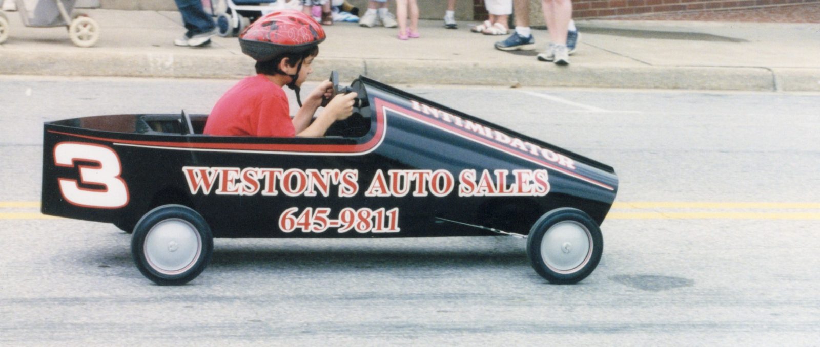 Young Chuck in a red helmet driving the black number 3 soap box car down the street.
