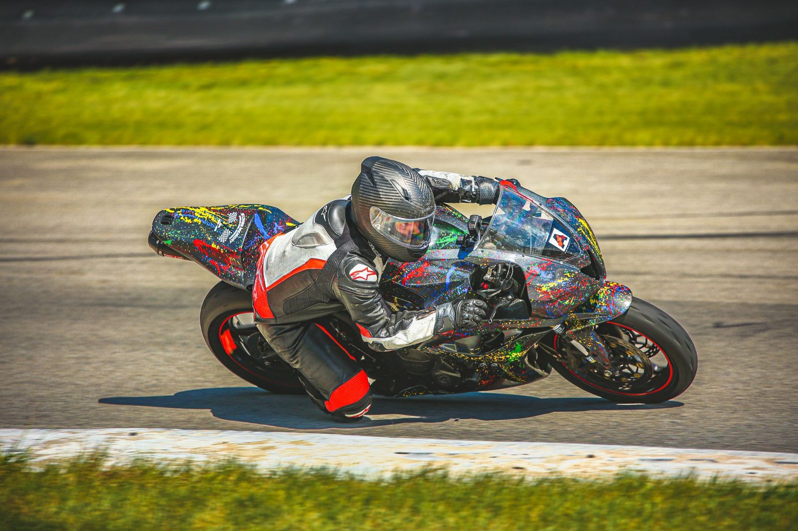 Chuck at full lean on the splatter-painted race bike, dark gray flecked with red, yellow, blue and green paint, knee down, bright green grass behind.